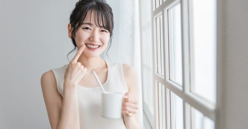 白いタンクトップの女性が窓際で歯を見せながら笑い、指で歯を指しながらコップを持っている様子